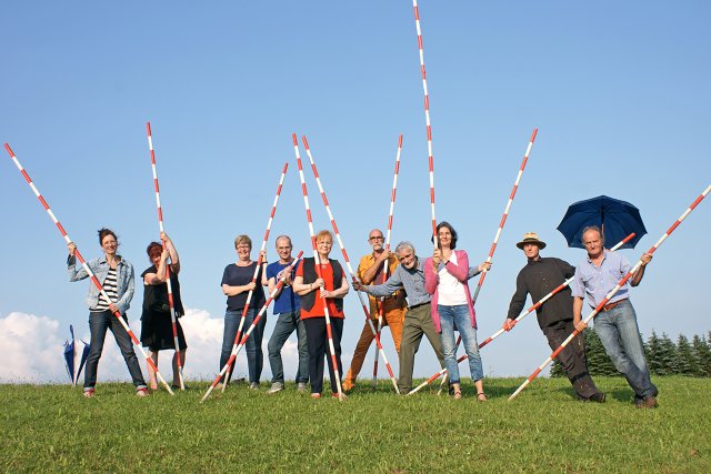 Das KunstWerkWege-Team mit langen rot-weiß gestreiften Stangen auf grüner Wiese vor blauem Himmel.