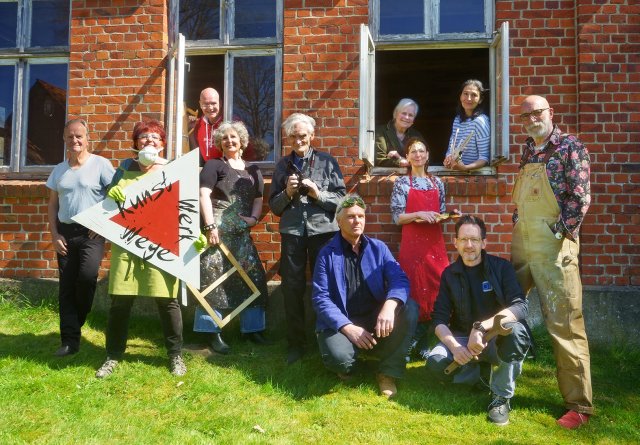 Das KunstWerkWege-Team vor einem roten Backsteingebäude mit geöffneten Fenstern, aus denen einige Mitglieder herausschauen.